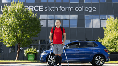 A young man is walking away from his blue vehicles using crutches. He had a red top on with a black backpack and blue shorts. He is looking away from the camera. Behind him are the words "sixth form college" in letters across a grey large building.
