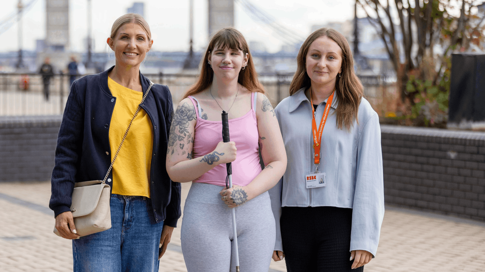 Lyndsey, Kaitlin and Rosie are photographed outside in London. Lyndsey is wearing a yellow top and navy jacket. Kaitlin is wearing a pink top and holding her white cane. Rosie is wearing a grey jumper. 