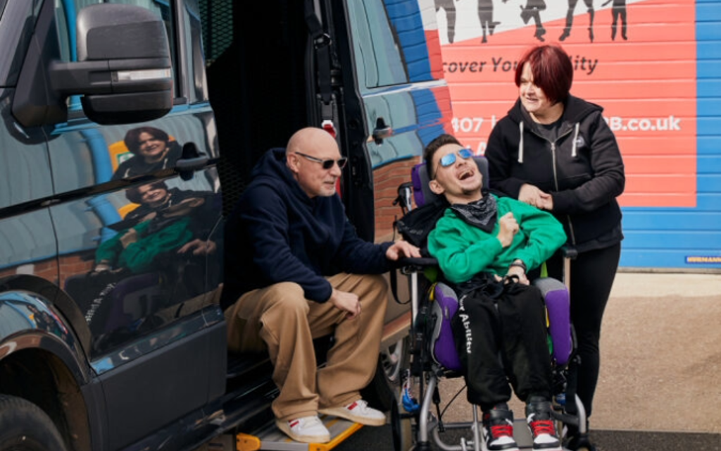 The photo is taken outside. A man is sitting on the step of the open door of a black vehicle to the left-hand side. In the middle of the photo is a young man smiling. He is sitting in his wheelchair. To the right-hand side of the image is a woman, she is looking at the man. Those photographed are Warren, Tyler and Teresa.