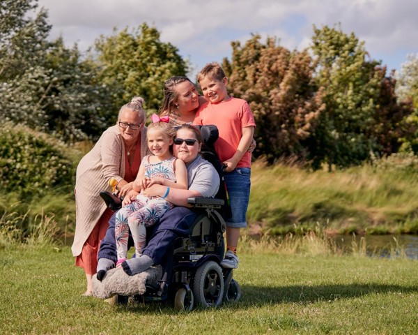 Spencer and his family are enjoying the sunny outdoor stole on a grassy field. They are smiling and hugging.
