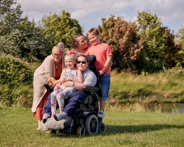 Spencer and his family are enjoying the sunny outdoor stole on a grassy field. They are smiling and hugging. 