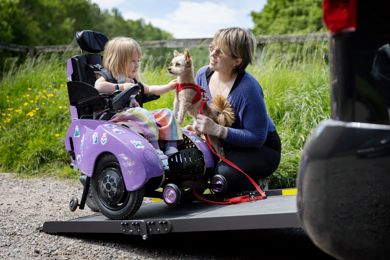 Poppy is sitting in her powered wheelchair on the ramp of her vehicle. Her mum Kim is crouched next to her, holding their family dog. Poppy is petting the dog and smiling.