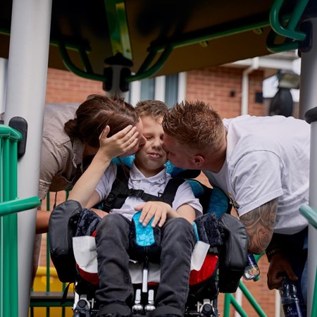 Lucas is sitting in his wheelchair. He is smiling. His mum Becci and dad Andy are kissing his checks on each side. 