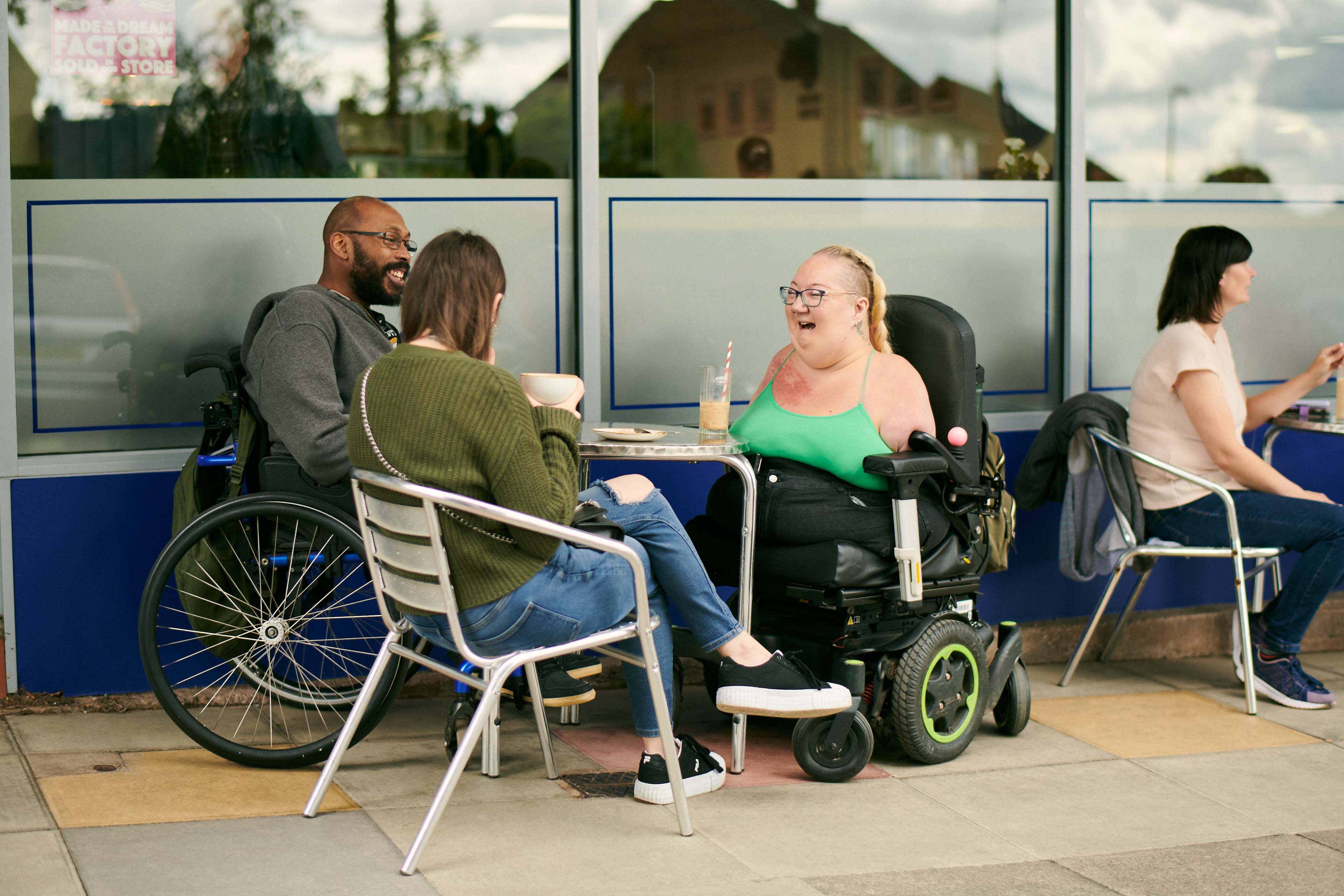 Tina is sitting with her friends laughing outside a café, they are sitting around a table. One is a lady sitting on a chair, holding a large mug. The other a gentleman in a powered wheelchair smiling.