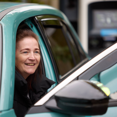Leanne is climbing out of the front seat of her blue vehicle smiling. She has long brown hair and black coat.