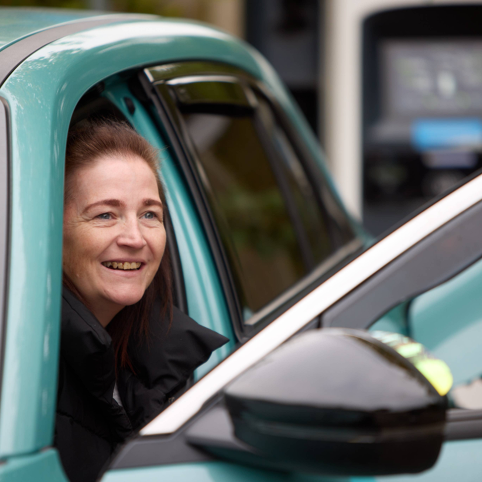 Leanne is climbing out of the front seat of her blue vehicle smiling. She has long brown hair and black coat.