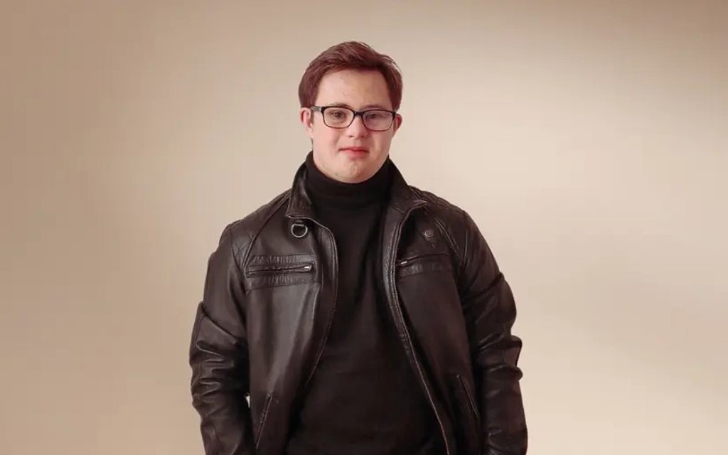 Ruben is standing facing the camera. He has brown short hair, black rimmed glasses, black leather jacket, black turtle neck and checkered grey and black trousers with a silver chain. 