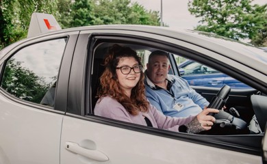 Imogen is sitting in the driving seat of her instructors car, it has a learner plate on top of the vehicle. She has light brown hair, black glasses, lilac jumper, black tattoos on her right hand and she is smiling. Her instructor is in the passenger seat. He has short dark hear and a light blue polo top on. 