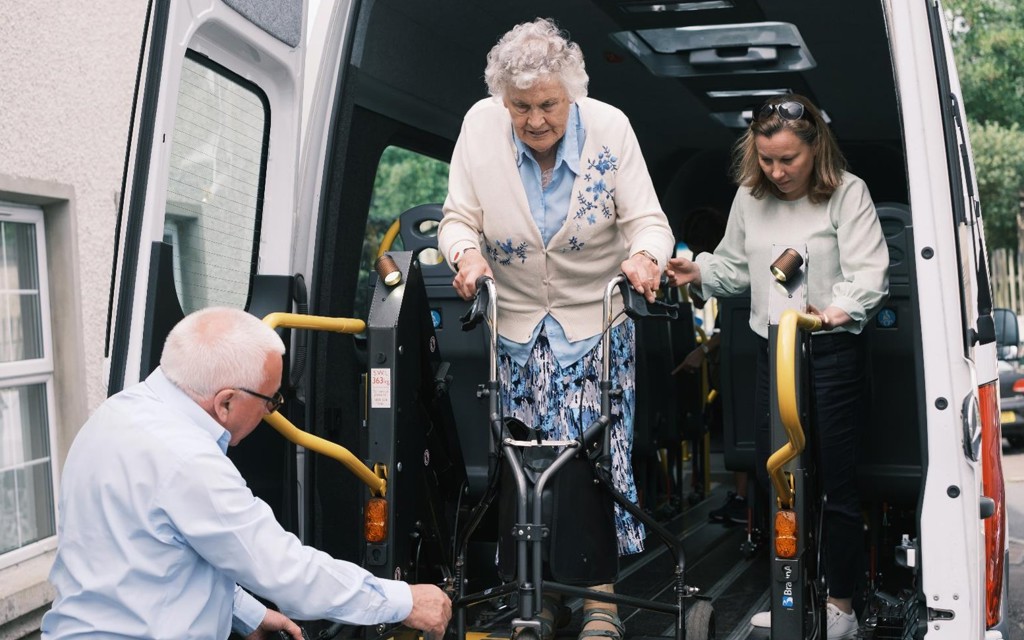 Lisa is standing in the back of a minibus with the doors open, she is supporting an older women who is stepping onto a lift at the back of the vehicle with her mobility aid. A gentleman is assisting the older women by controlling the lift. 