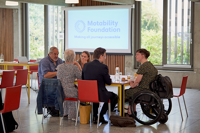 A group of people discussing issues at a table. A Motability Foundation logo sits in the background.