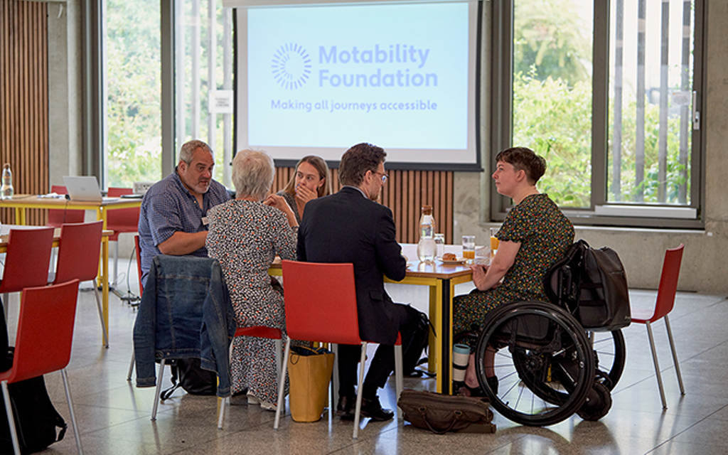 A group of people discussing issues at a table. A Motability Foundation logo sits in the background.
