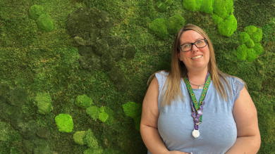 This image shows Danielle smiling at the camera stood in front of a green moss covered wall. She has long brown hair and is wearing black-framed glasses and a light blue top. Around her neck is a silver necklace, her purple work lanyard and her green sunflower lanyard. Her lanyards are decorated with various purple pin badges.