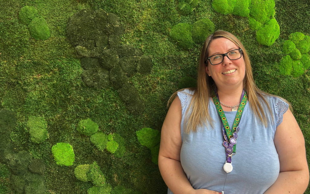 This image shows Danielle smiling at the camera stood in front of a green moss covered wall. She has long brown hair and is wearing black-framed glasses and a light blue top. Around her neck is a silver necklace, her purple work lanyard and her green sunflower lanyard. Her lanyards are decorated with various purple pin badges.