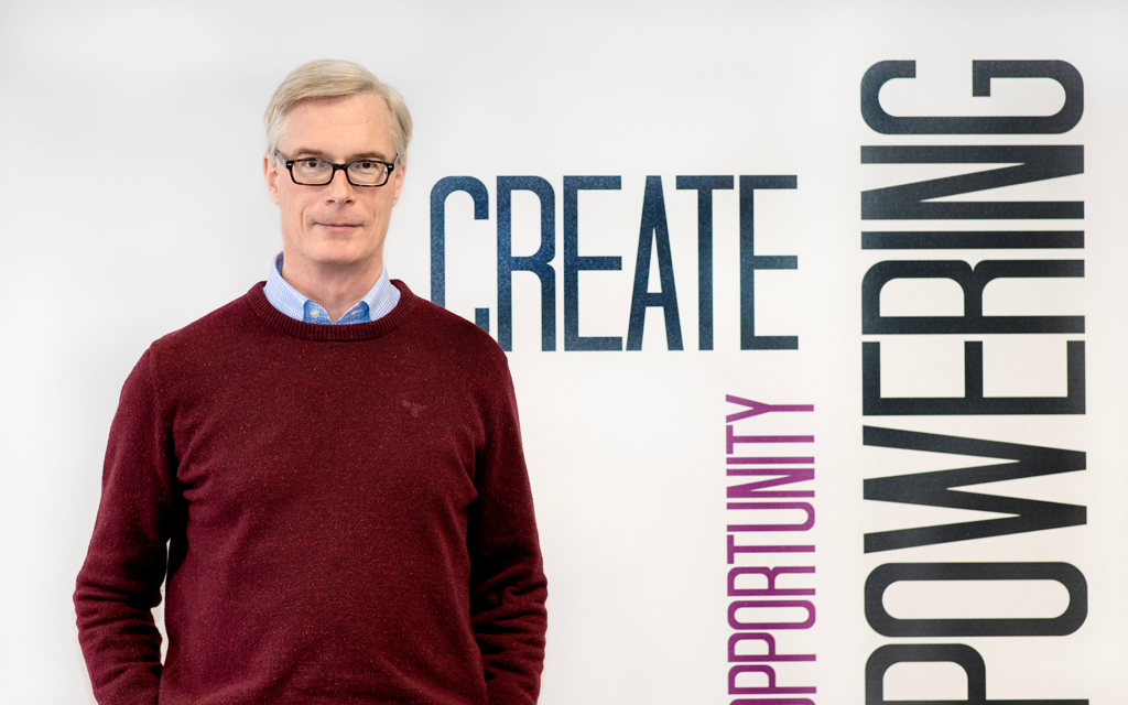 Charles is smiling and standing in front of a colourful background.
