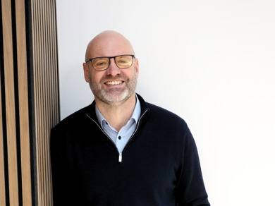 Nigel Fletcher CEO is smiling in a black jumper, blue shirt with black rimmed glasses. 