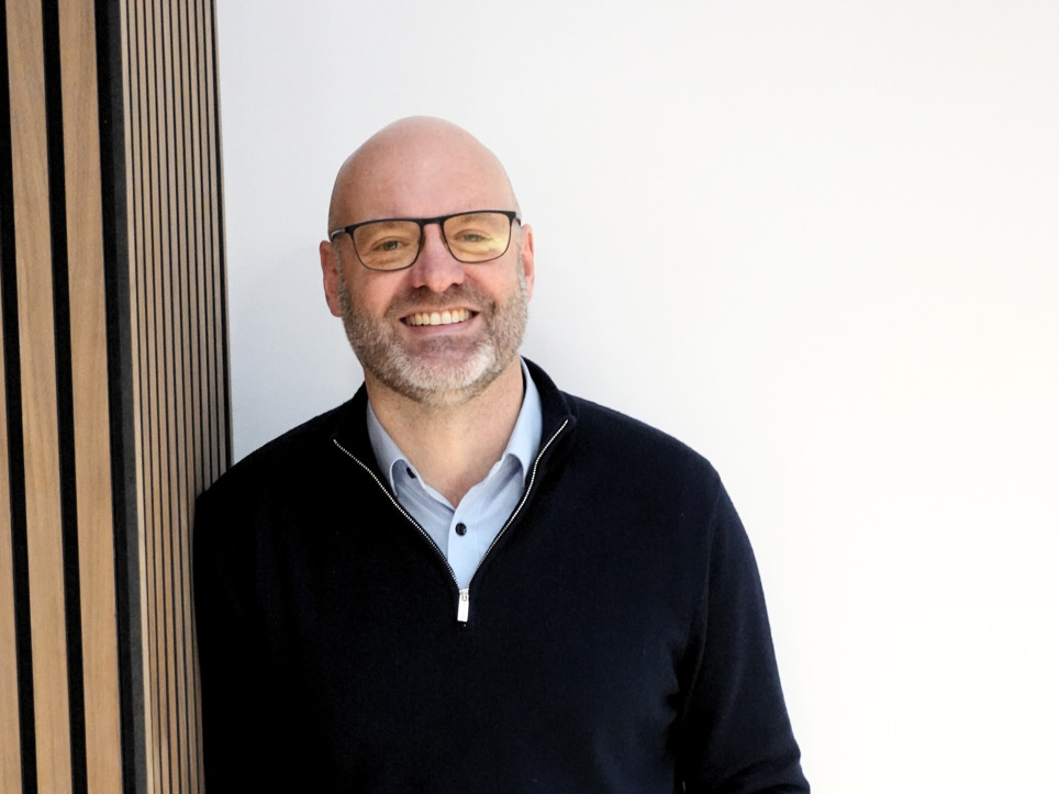 Nigel Fletcher CEO is smiling in a black jumper, blue shirt with black rimmed glasses. 