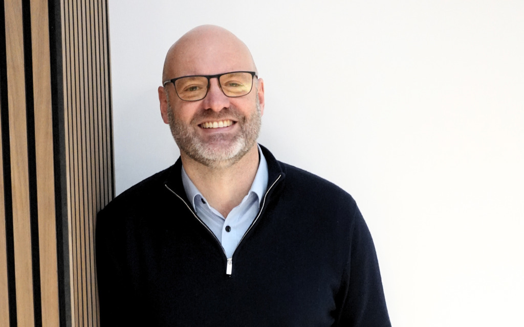 Nigel Fletcher CEO is smiling in a black jumper, blue shirt with black rimmed glasses. 