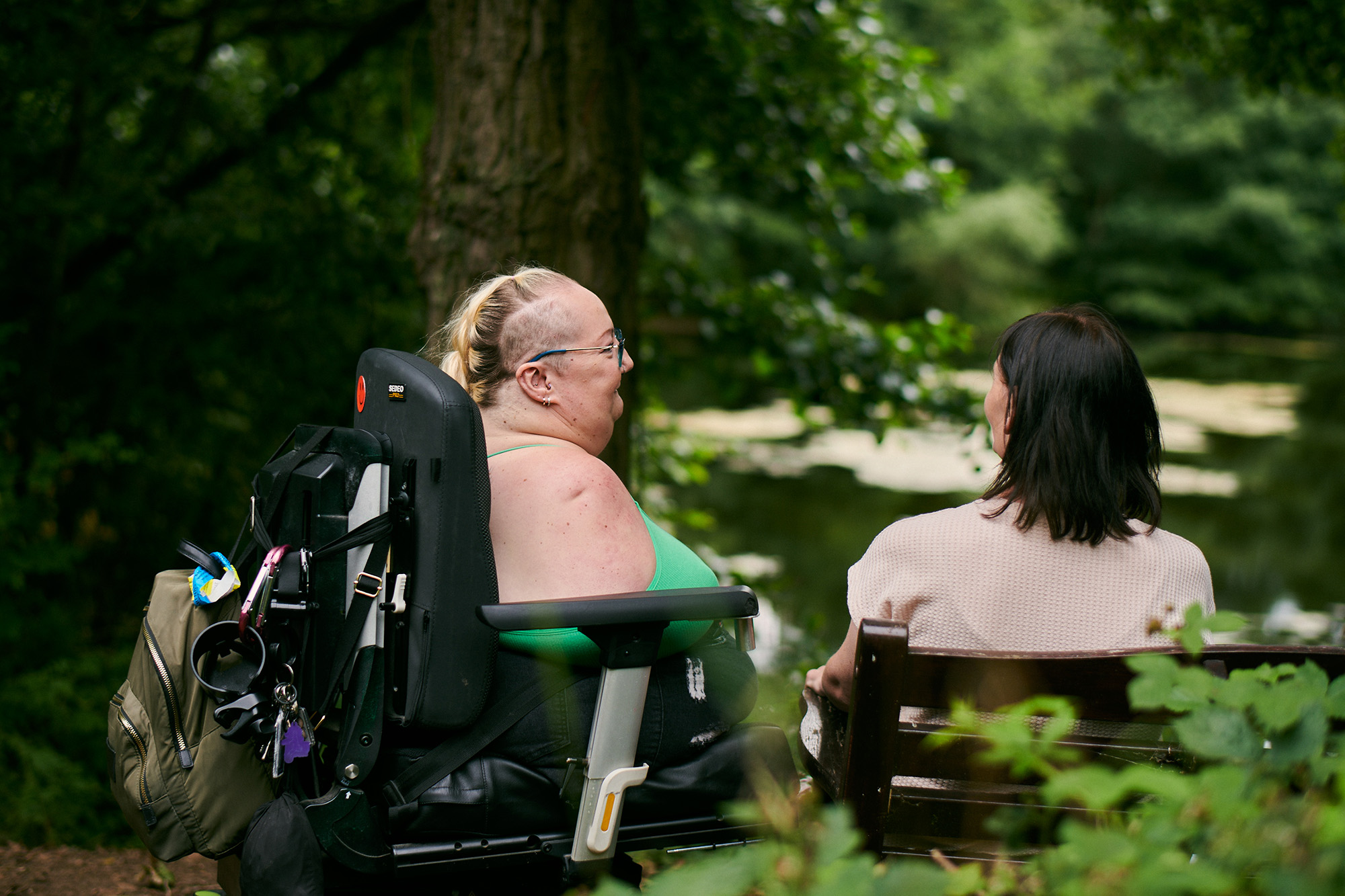 Tina and her friend are sitting together looking over a lake in a peaceful park. 