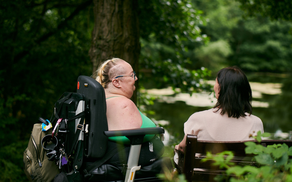 Tina and her friend are sitting together looking over a lake in a peaceful park.