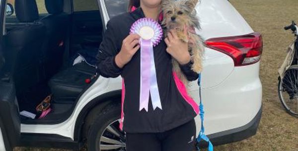 A young women is smiling holding a small dog in one hand and a rosette in another.