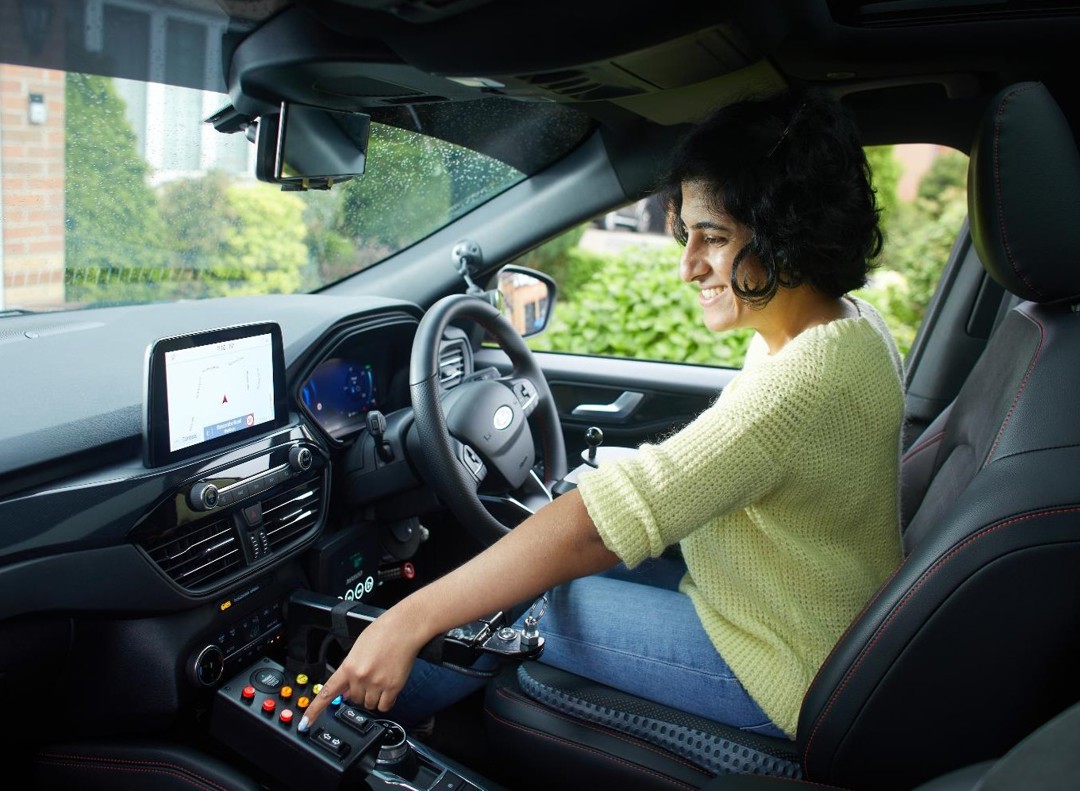 Mathy is sitting in the drivers seat of her vehicle smiling. She is looking to the left and choosing a button on a panel within her vehicle. She is wearing a yellow knitted jumper, blue jeans and has short black hair in a bob.