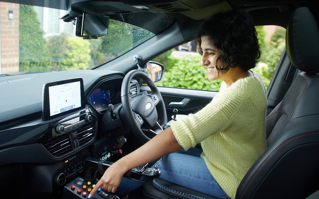 Mathy is sitting in the drivers seat of her vehicle smiling. She is looking to the left and choosing a button on a panel within her vehicle. She is wearing a yellow knitted jumper, blue jeans and has short black hair in a bob.