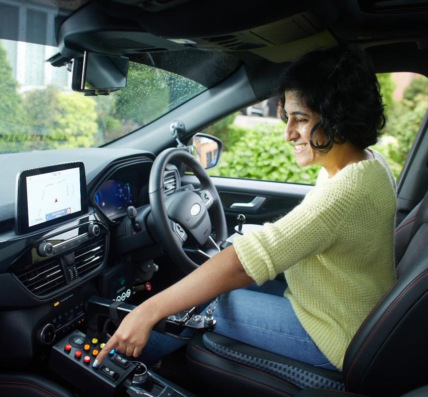 Mathy is sitting in the drivers seat of her vehicle smiling. She is looking to the left and choosing a button on a panel within her vehicle. She is wearing a yellow knitted jumper, blue jeans and has short black hair in a bob.