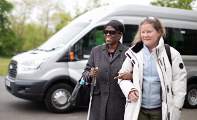 A women has linked arms with another woman as they walk away from a silver minibus which sits in the background. The first women is smiling, she is wearing a wool coat, sun glasses, black handbag and is using a walking aid. The second women has a white coat on and a light blue fleece underneath with sunglasses on her head. 