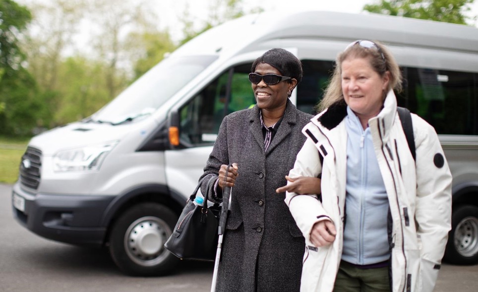 A women has linked arms with another woman as they walk away from a silver minibus which sits in the background. The first women is smiling, she is wearing a wool coat, sun glasses, black handbag and is using a walking aid. The second women has a white coat on and a light blue fleece underneath with sunglasses on her head. 