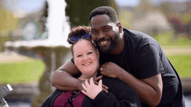 Steph is sat on her mobility scooter, holding her walking aid. She is wearing a burgundy top with black ribbon bows and a black knitted cardigan. She has her hair in a bun with sunglasses resting on her head in front of her bun. She is being hugged by her husband David who is wearing a black t shirt. They are both smiling at the camera.