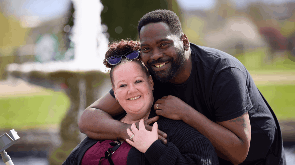 Steph is sat on her mobility scooter, holding her walking aid. She is wearing a burgundy top with black ribbon bows and a black knitted cardigan. She has her hair in a bun with sunglasses resting on her head in front of her bun. She is being hugged by her husband David who is wearing a black t shirt. They are both smiling at the camera.