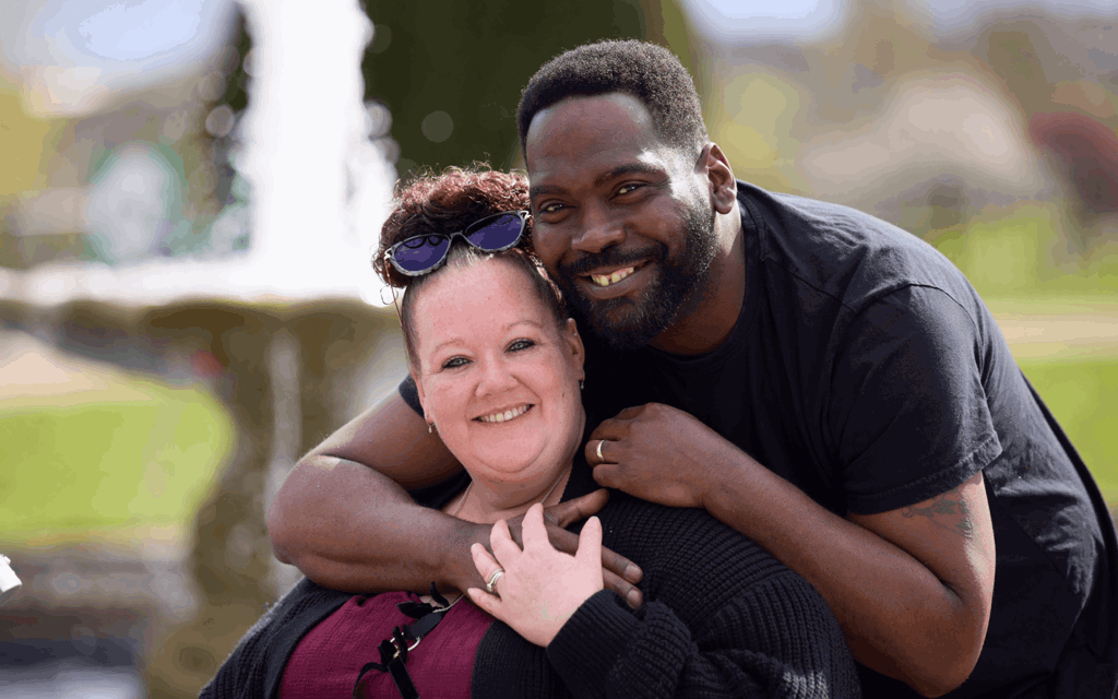 Steph is sat on her mobility scooter, holding her walking aid. She is wearing a burgundy top with black ribbon bows and a black knitted cardigan. She has her hair in a bun with sunglasses resting on her head in front of her bun. She is being hugged by her husband David who is wearing a black t shirt. They are both smiling at the camera.
