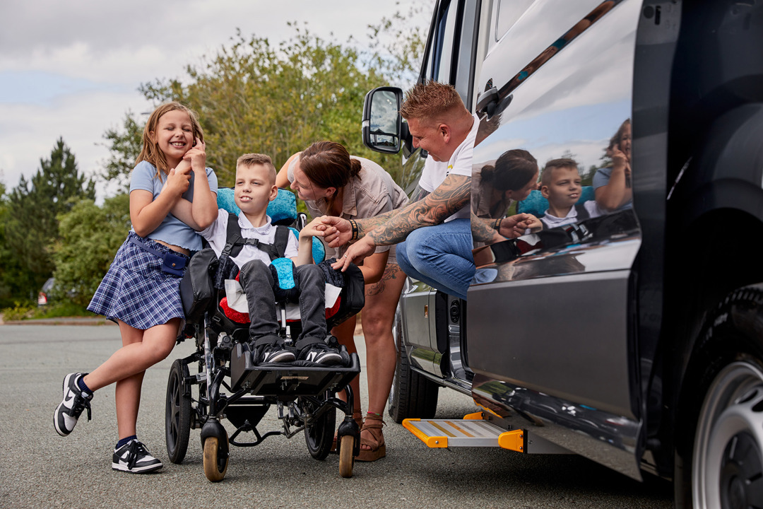 Lucas is in his wheelchair next to his black vehicle. He is smiling. His sister is standing to the left of him smiling and holding his hand. His mum Becci is standing to the right of Lucas and is talking to him. His dad Andy is to the right of Becci and is sitting in the vehicle with the door open smiling at Lucas.