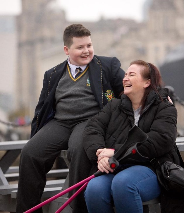 Leanne is sitting on a picnic bench laughing, she has long brown hair, black coat, black handbag, blue jeans and black and pink crutches. Sitting next to her is her son. He has short brown hair and is wearing his school uniform smiling.