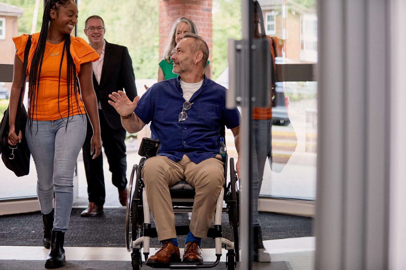 Colleagues are walking into their office building. A lady is laughing and chatting to a gentleman in a wheelchair. 