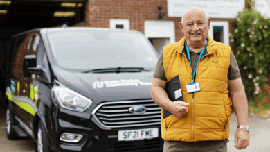 The photograph is of a man standing in from of a black community transport vehicle. The man is wearing a yellow gilet with a green polo shirt. He is holding a clipboard and is smiling to camera.