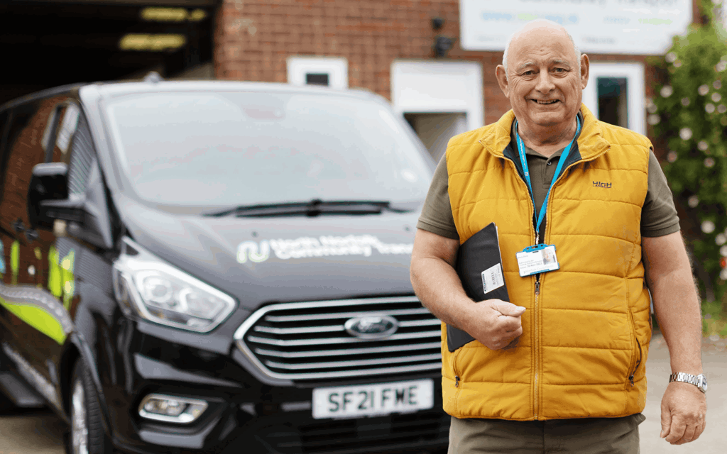 The photograph is of a man standing in from of a black community transport vehicle. The man is wearing a yellow gilet with a green polo shirt. He is holding a clipboard and is smiling to camera.