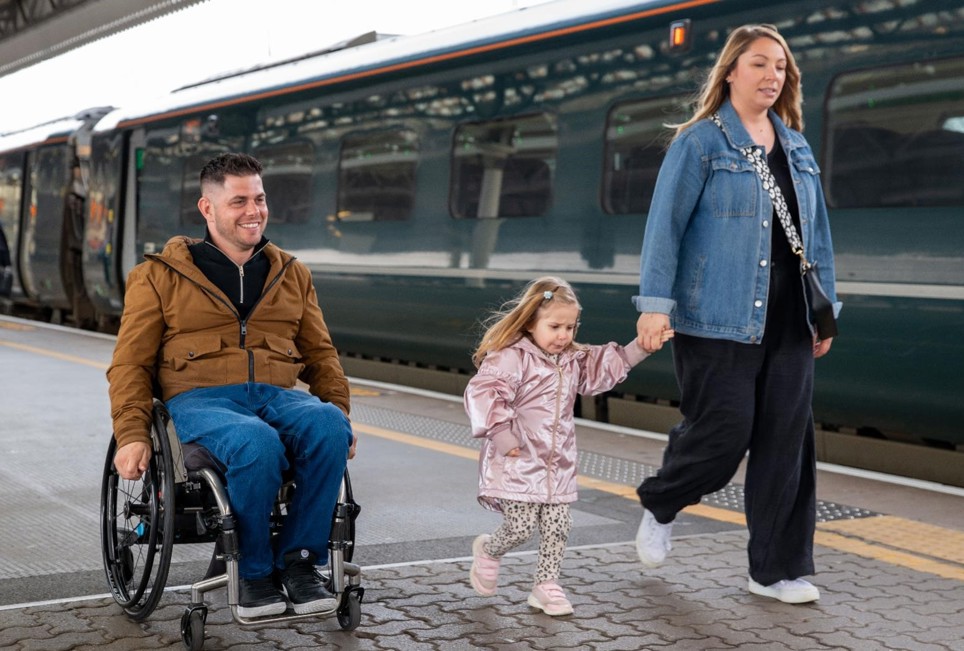 The image shows a man in a wheelchair on a train station platform, along with a woman and a young girl. The man is wearing a brown coat, black jumper, blue jeans, and black trainers. He wheeling along the platform.  Accompanying the man is a woman in a denim jacket, black jumpsuit, and white trainers. She is walking and holding hands with a young girl. The girl is dressed in a pink raincoat, white and black leopard print leggings, and pink trainers. A train is visible in the station next to the group. 