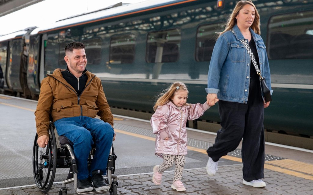 The image shows a man in a wheelchair on a train station platform, along with a woman and a young girl. The man is wearing a brown coat, black jumper, blue jeans, and black trainers. He wheeling along the platform.  Accompanying the man is a woman in a denim jacket, black jumpsuit, and white trainers. She is walking and holding hands with a young girl. The girl is dressed in a pink raincoat, white and black leopard print leggings, and pink trainers. A train is visible in the station next to the group. 