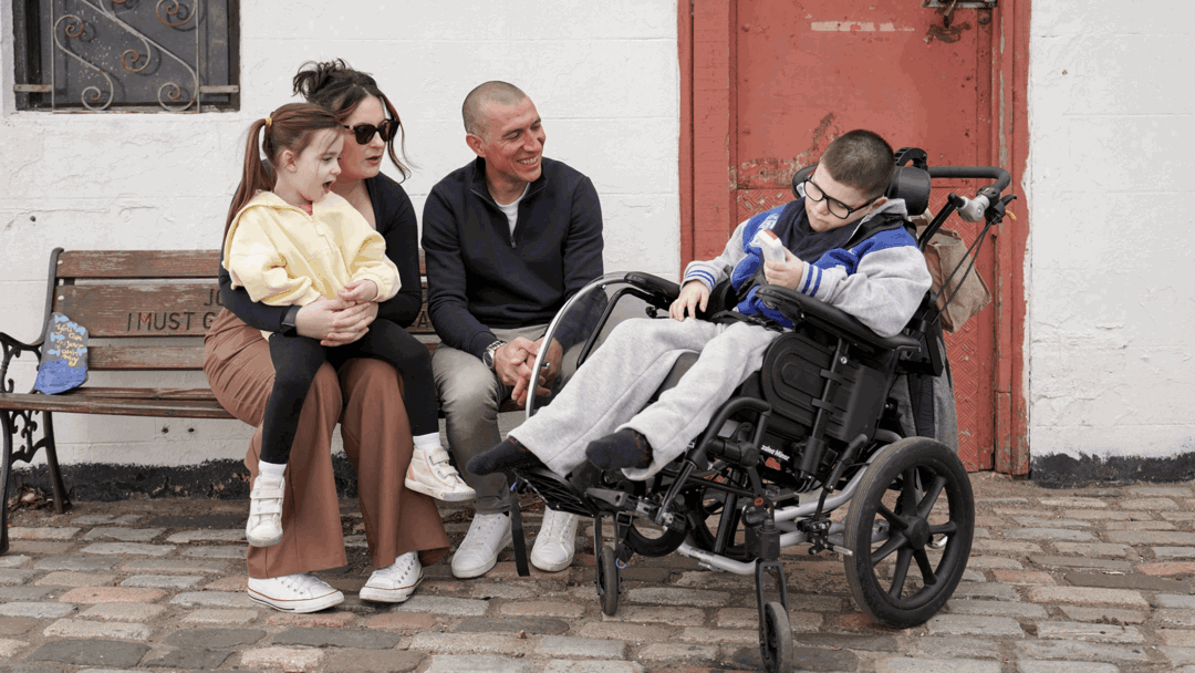 Gino is sat in his wheelchair next to his mum, sister and dad, who are sat on a bench. They are laughing and talking outside. His sister is sat on his mums lap. 