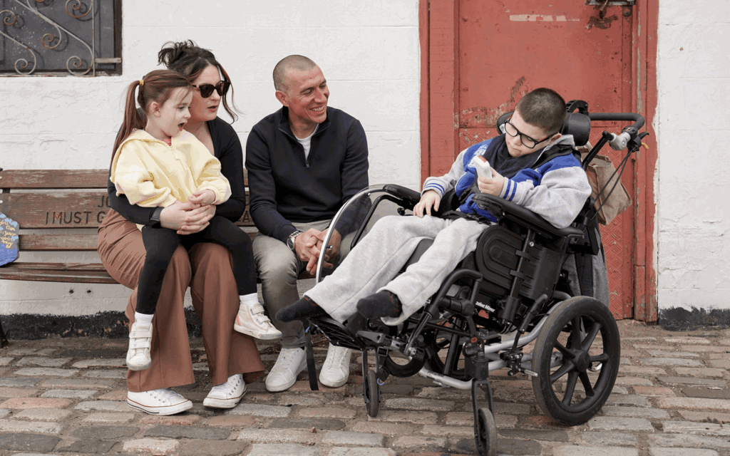 Gino is sat in his wheelchair next to his mum, sister and dad, who are sat on a bench. They are laughing and talking outside. His sister is sat on his mums lap. 