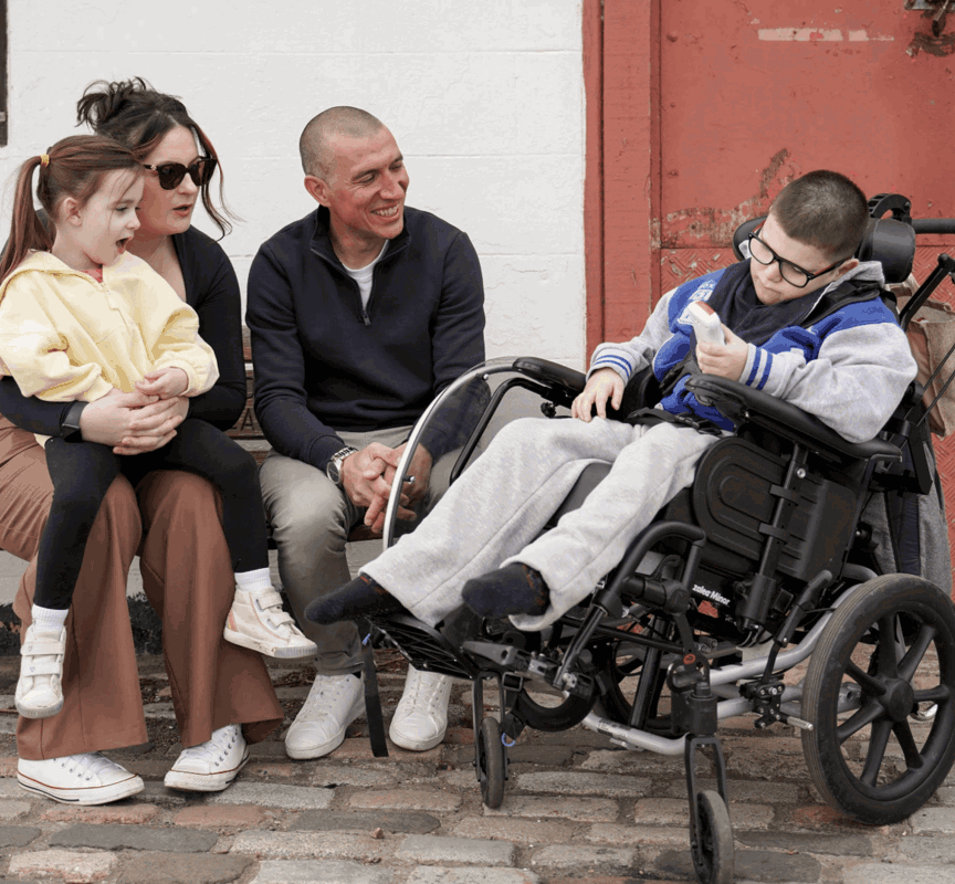 Gino is sat in his wheelchair next to his mum, sister and dad, who are sat on a bench. They are laughing and talking outside. His sister is sat on his mums lap. 