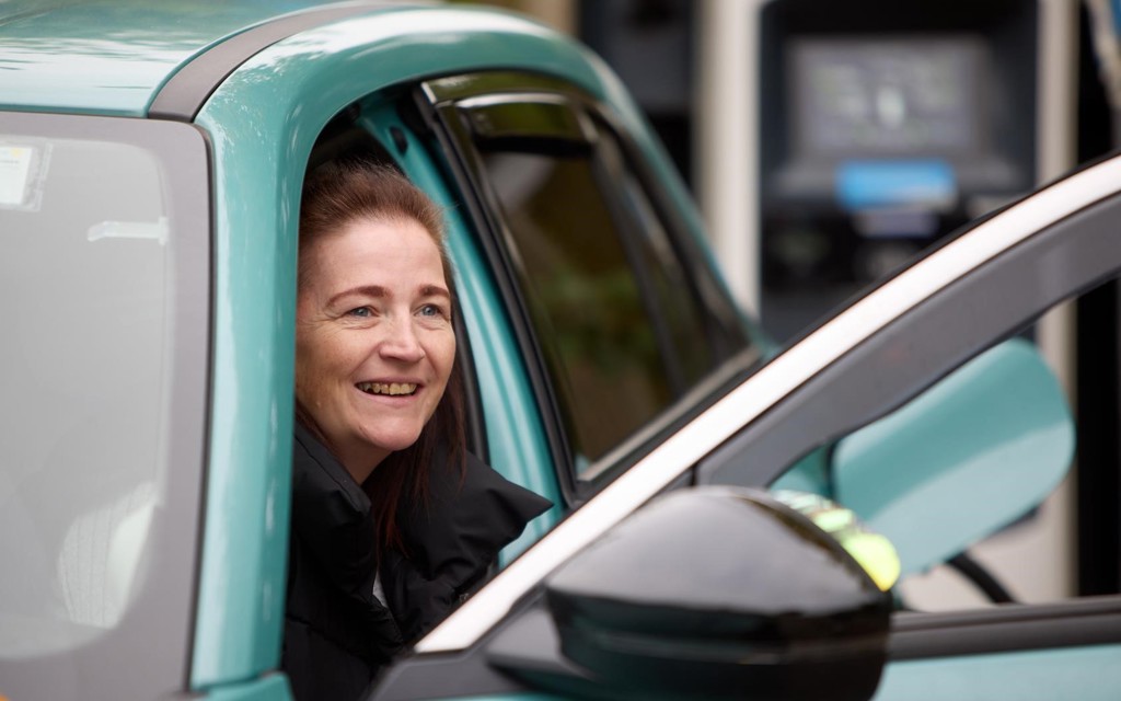 Leanne is climbing out of the front seat of her blue vehicle smiling. She has long brown hair and black coat.