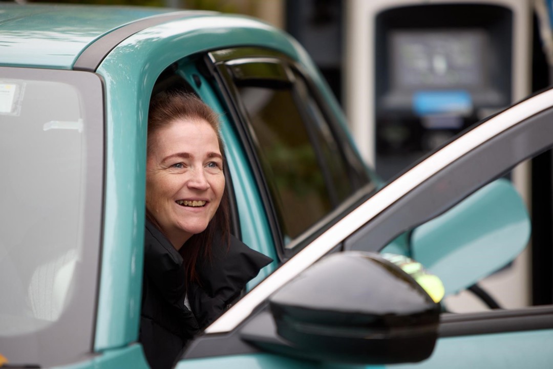 Leanne is climbing out of the front seat of her blue vehicle smiling. She has long brown hair and black coat.