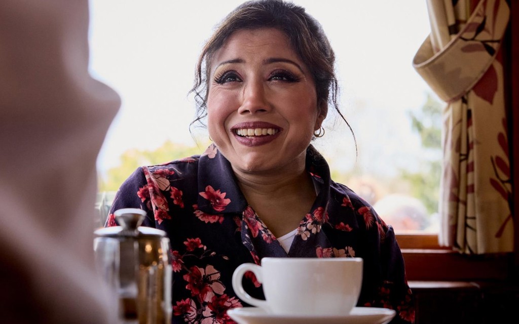 Nazmin is sitting at a table in a café with a cup of tea. She is smiling brightly while talking to a friend. She has brown hair tied in a bun and is wearing a navy shirt with pink flowers. 