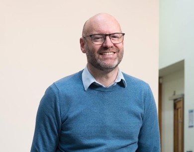 Nigel Fletcher CEO is smiling in a blue jumper with black rimmed glasses. 
