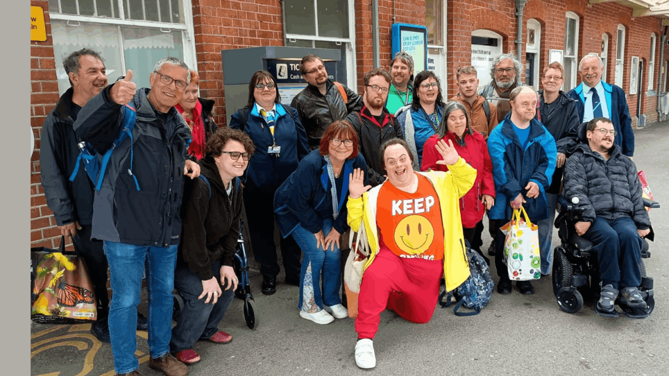 The photo is of a large group of 18, non-disabled people and disabled people, on a train platform. Everyone is looking at the camera smiling or waving.