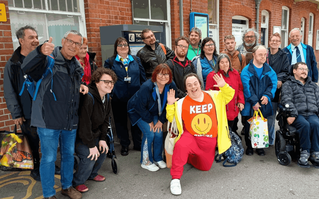 The photo is of a large group of 18, non-disabled people and disabled people, on a train platform. Everyone is looking at the camera smiling or waving.