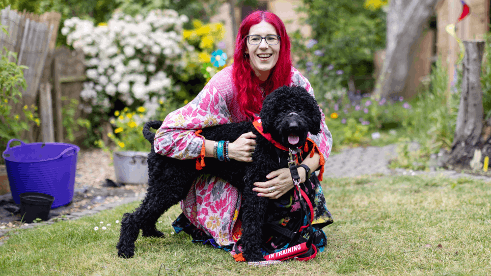 Charlie has pink hair and is wearing multi coloured clothing. They are holding their dog by the lead in their garden. Their dog is black poodle cross and is wearing a red neckerchief with a "in training" sign on their lead.