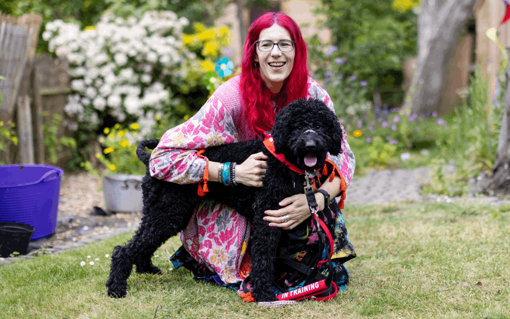 Charlie has pink hair and is wearing multi coloured clothing. They are holding their dog by the lead in their garden. Their dog is black poodle cross and is wearing a red neckerchief with a "in training" sign on their lead.